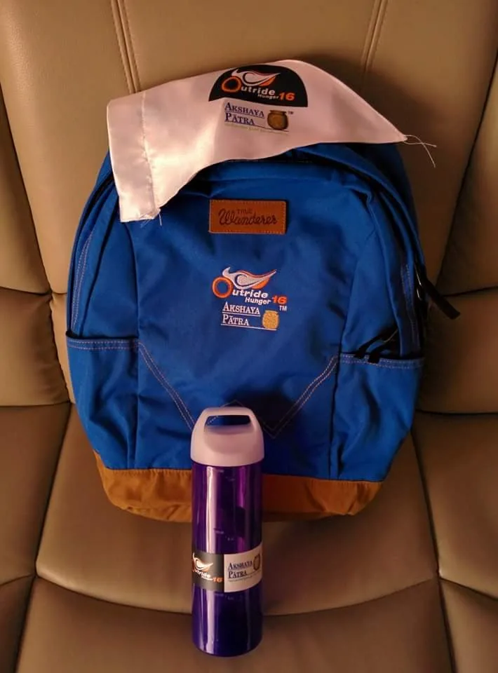 Blue backpack embroidered with the Akshaya Patra Outride Hunger logo, shown with a branded bottle and printed cloth